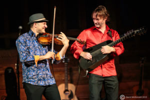 Two White men play violin and flamenco guitar while looking at each other. / Deux hommes Blancs jouent du violon et de la guitare flamenca en se regardant.