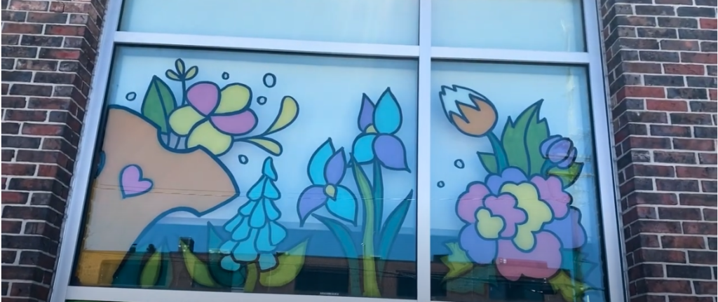 A large window with fun cartoon style flowers that are pink, yellow, orange, white, purple, green, and all are outlined in black. It is fun, uplifting, and one of the images is an orange t shirt with a pink heart.