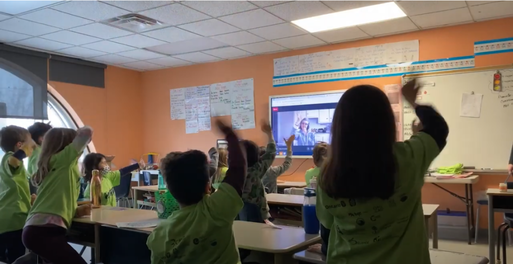 A group of elementary school aged children in a classroom with one of their hands up in the air. Most are wearing a green t-shirt and they are watching a video on a projected screen.