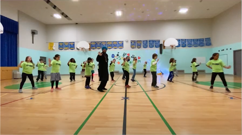 A group of elementary school aged children spread out in a gymnasium with one adult in the front. They all have one of their arms out in front of them as part of a dance routine.