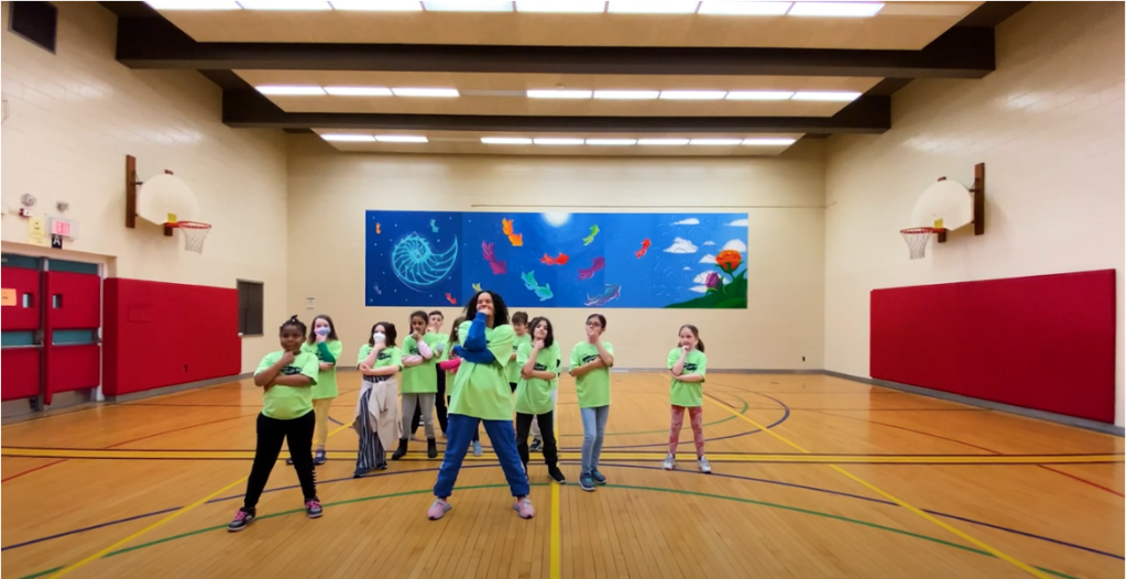 A group of elementary school aged children and one adult in front of them, all in green t shirts. They are posing with one hand under their chin and another tucked under their elbows as part of a dance routine.