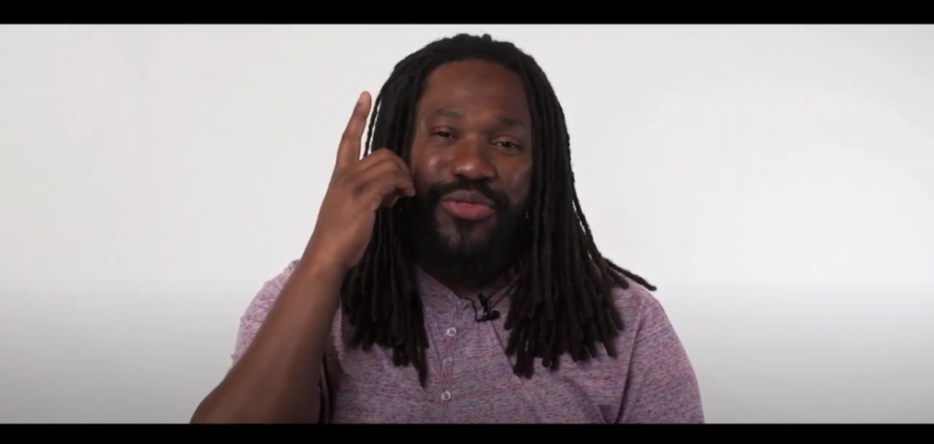 JustJamaal ThePoet pointing upwards in the center of a white background. He has a black short beard, black dreads, and is wearing a purple buttoned up t shirt.