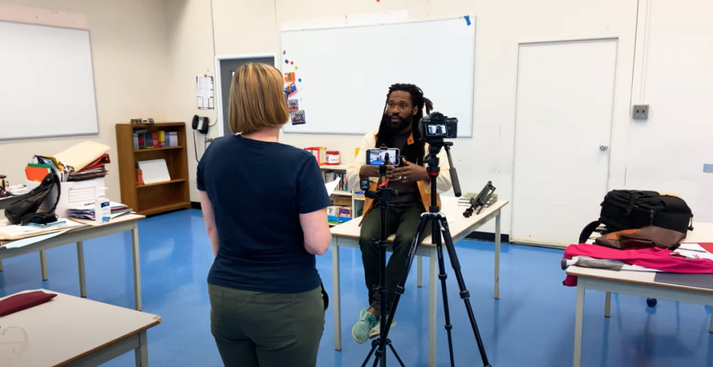 A woman with short dark blond hair standing in a class room behind a camera as well as a phone. She is filming JustJamaal ThePoet who is sitting on a table talking.