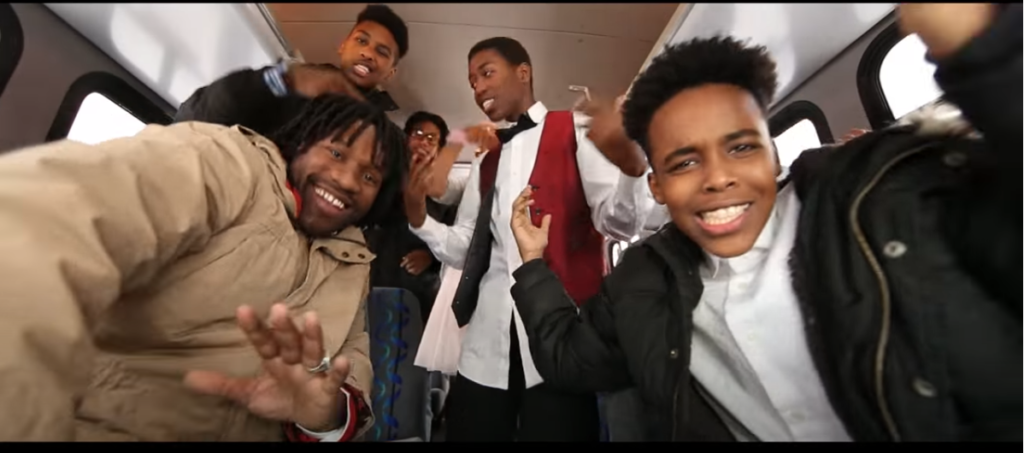 A group of young men, some in large jackets and one of them in a bow time and red vest, looking into a camera. It is a screenshot from Just Jamaal the Poet's music video.