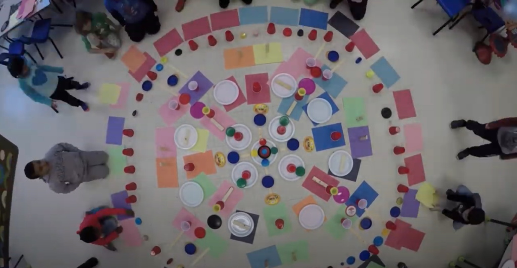 A birds eye view of circular pattern of blues, pinks, green, orange, and purple construction paper and paper plates, cups, and lids, with children surrounding the art.