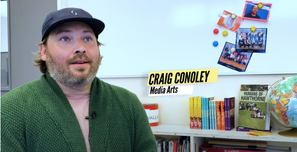 Craig Conoley, a media artist, in a green cardigan with a grey cap sitting in front of a bookshelf with a few class photos behind him. He is being interviewed.