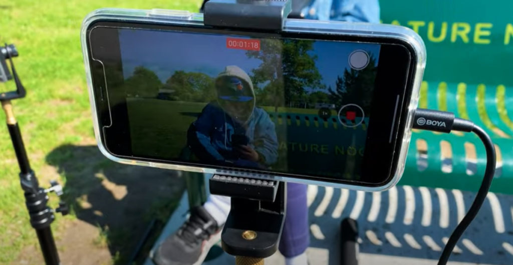 A shot of a phone that is taking a video of a man in a Blue Jays cap, a white hoodie, and blue jacket sitting on a bench. The man is looking at his phone on a sunny day in a field.