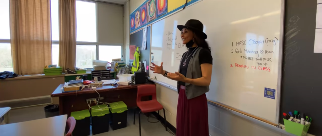 Jacqui-Du-Toit is standing in a classroom in front of a white board with both her arm up, an eager face, looking passionate about what she is teaching.