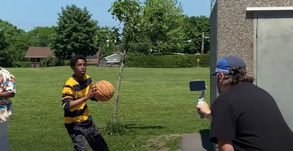 A young man holding a basket ball looking at where he will be aiming to throw the ball. There is a middle aged man filming him on his phone for a video and they are in a sunny field.