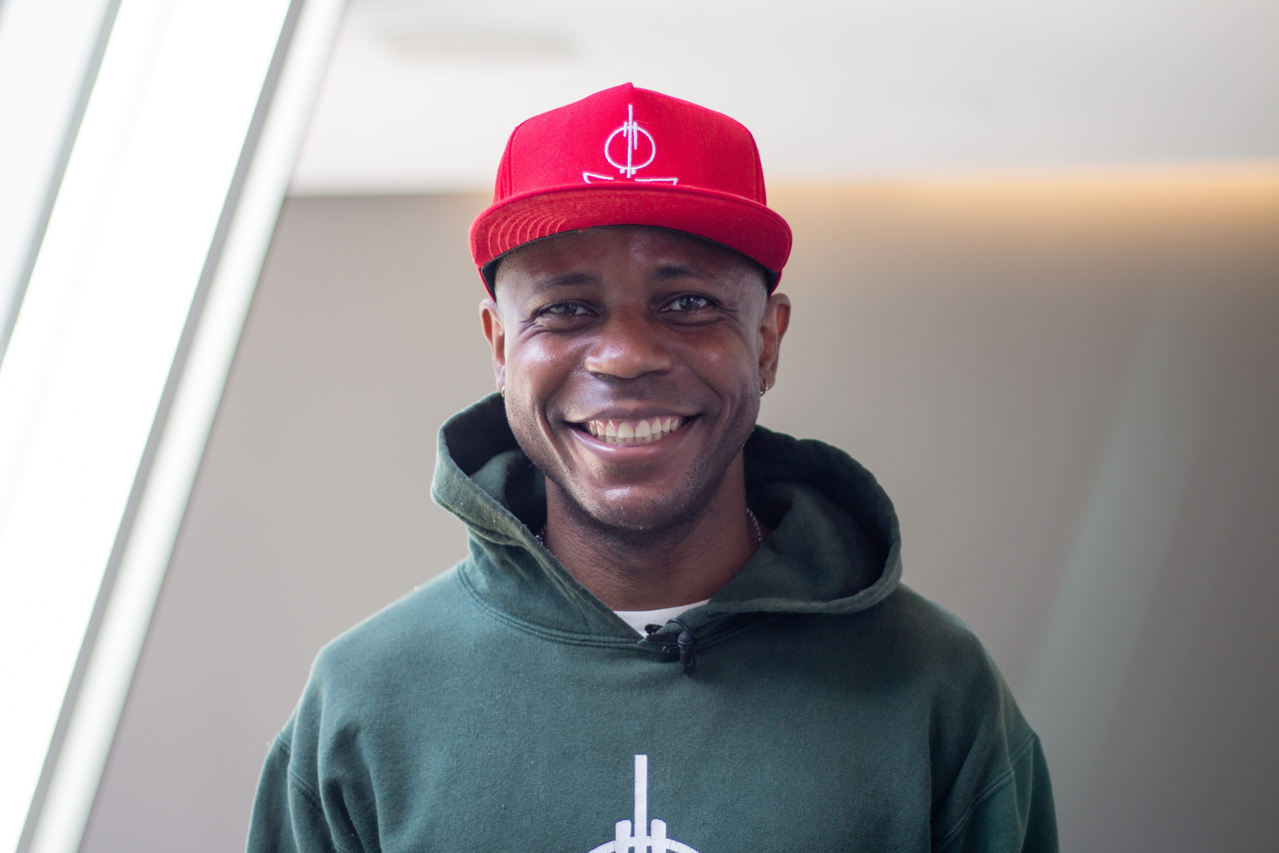 A Black man wearing a red baseball cap and dark green hoodie smiles at the camera. / Un homme Noir en casquette rouge et pull vert foncé sourit à la caméra.