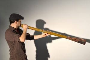 A smiling bearded White man plays the didgeridoo. / Un homme Blanc souriant et barbu joue du didgeridoo.