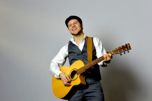 A smiling bearded White man plays the acoustic guitar. / Un homme Blanc souriant et barbu joue de la guitare acoustique.
