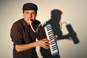 A smiling bearded White man plays the harmonium in front of a white backdrop, his silhouette behind him. / Un homme Blanc barbu souriant joue de l’harmonium devant un arrière-plan blanc, sa silhouette derrière lui.
