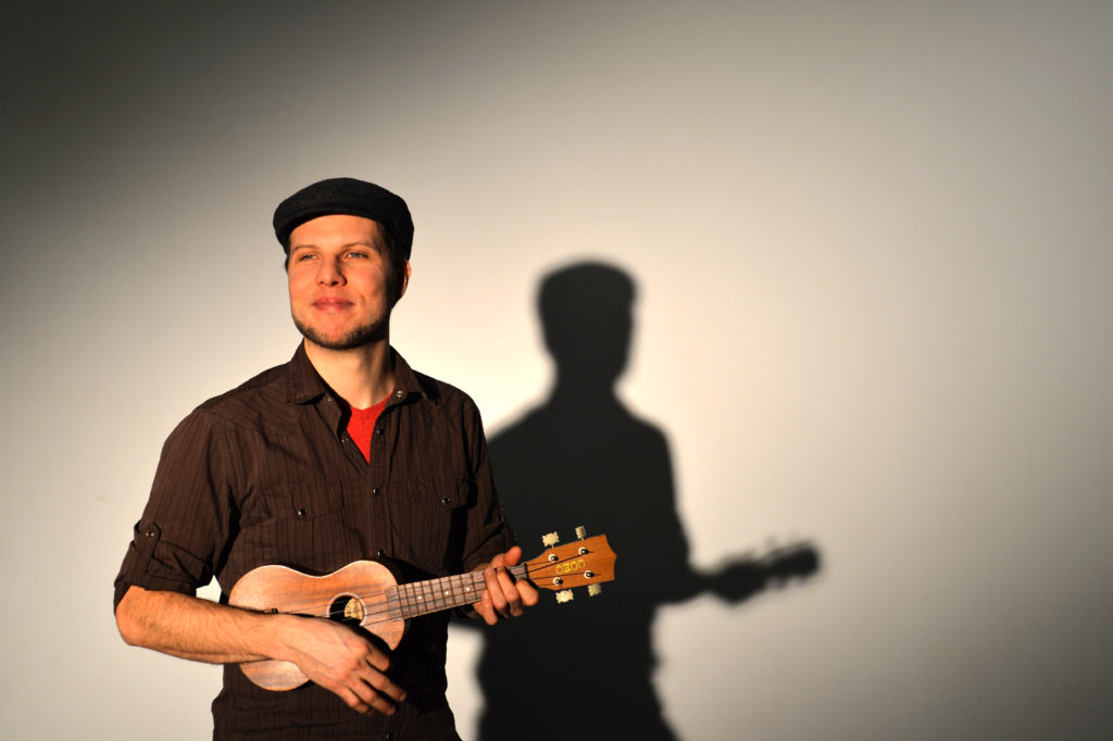A smiling bearded White man plays the ukulele in front of a white backdrop, his silhouette behind him. / Un homme Blanc barbu souriant joue de l’ukulele devant un arrière-plan blanc, sa silhouette derrière lui.