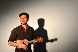 A smiling bearded White man plays the ukulele in front of a white backdrop, his silhouette behind him. / Un homme Blanc barbu souriant joue de l’ukulele devant un arrière-plan blanc, sa silhouette derrière lui.
