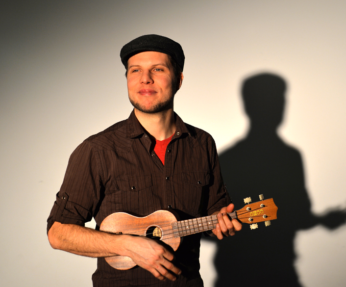A smiling bearded White man plays the ukulele in front of a white backdrop, his silhouette behind him. / Un homme Blanc barbu souriant joue de l’ukulele devant un arrière-plan blanc, sa silhouette derrière lui.