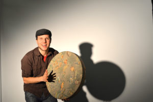 A smiling bearded White man plays a drum. / Un homme Blanc souriant et barbu joue du tambour.