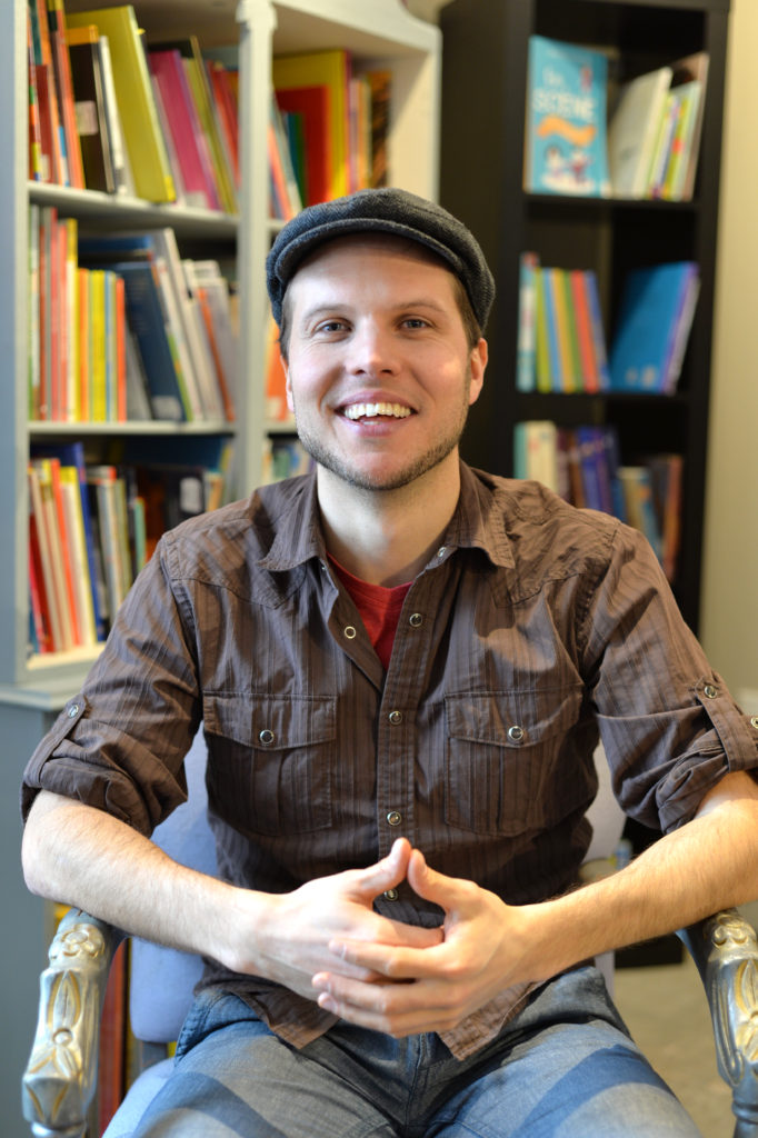 A bearded White man sits in a library and smiles at the camera. / Un homme Blanc barbu sourit à la caméra, assis dans une bibliothèque.
