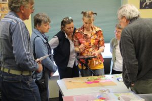 Five seniors stand around a White woman demonstrating something on her iPad, held up by a tripod, overlooking a collage. / Cinq séniors entourent une femme Blanche qui leur montre quelque chose sur son iPad, tenu au dessus d’un collage par un trépied.