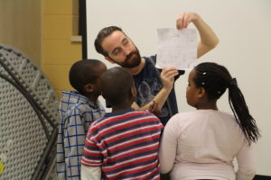 A White man holds up a piece of paper with pencil drawings on it for three Black children. / Un homme Blanc tient une feuille avec des dessins au crayon en l’air pour trois enfants Noirs.