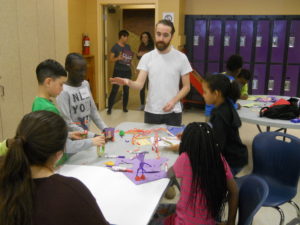 A White man talks to a table of children who are creating things with paper, tape, markers and chenille stems. / Un homme Blanc parle à une table d’enfants qui créent avec du papier, du scotch, des feutres et des tiges de chenille.