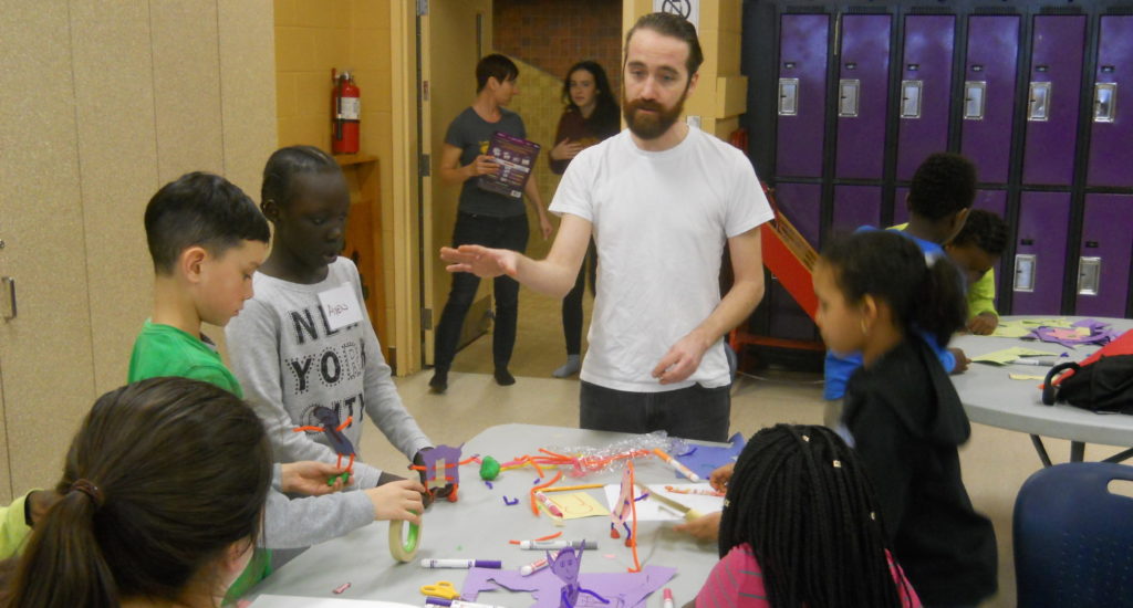 A White man talks to a table of children who are creating things with paper, tape, markers and chenille stems. / Un homme Blanc parle à une table d’enfants qui créent avec du papier, du scotch, des feutres et des tiges de chenille.