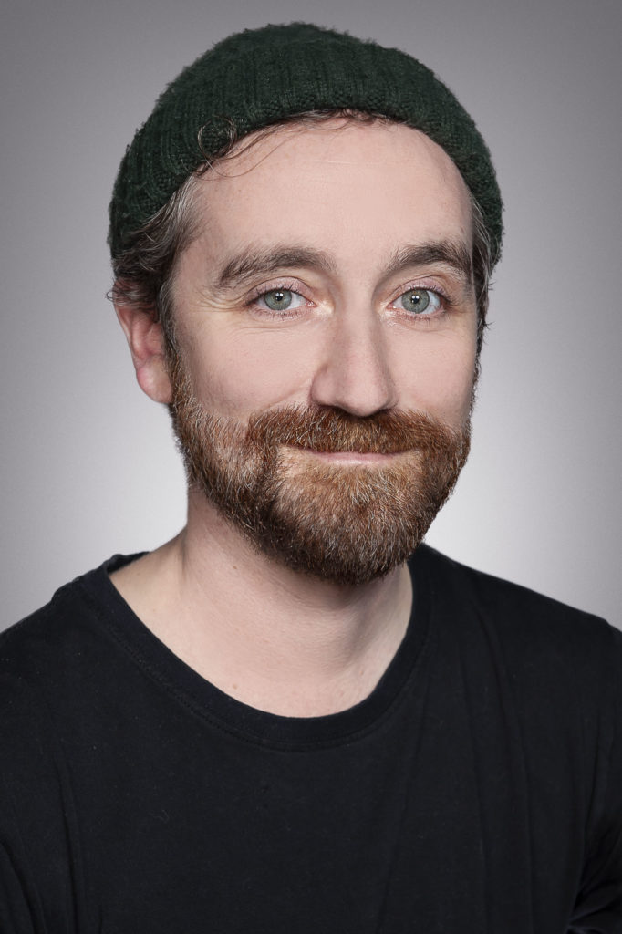 Headshot of a White bearded man in a beanie smiling at the camera. / Un gros plan d’un homme Blanc barbu qui porte un bonnet et sourit à la caméra.