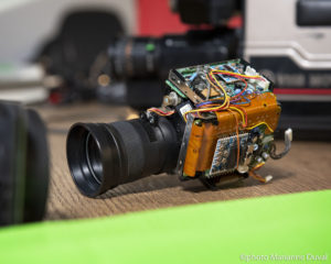 Close-up of a camera lens attached to exposed internal camera parts like wires, motherboards, and chips. / Gros plan de l’objectif d’un appareil photo attaché à des éléments internes exposés de l’appareil comme des cables, des cartes mères et des puces.