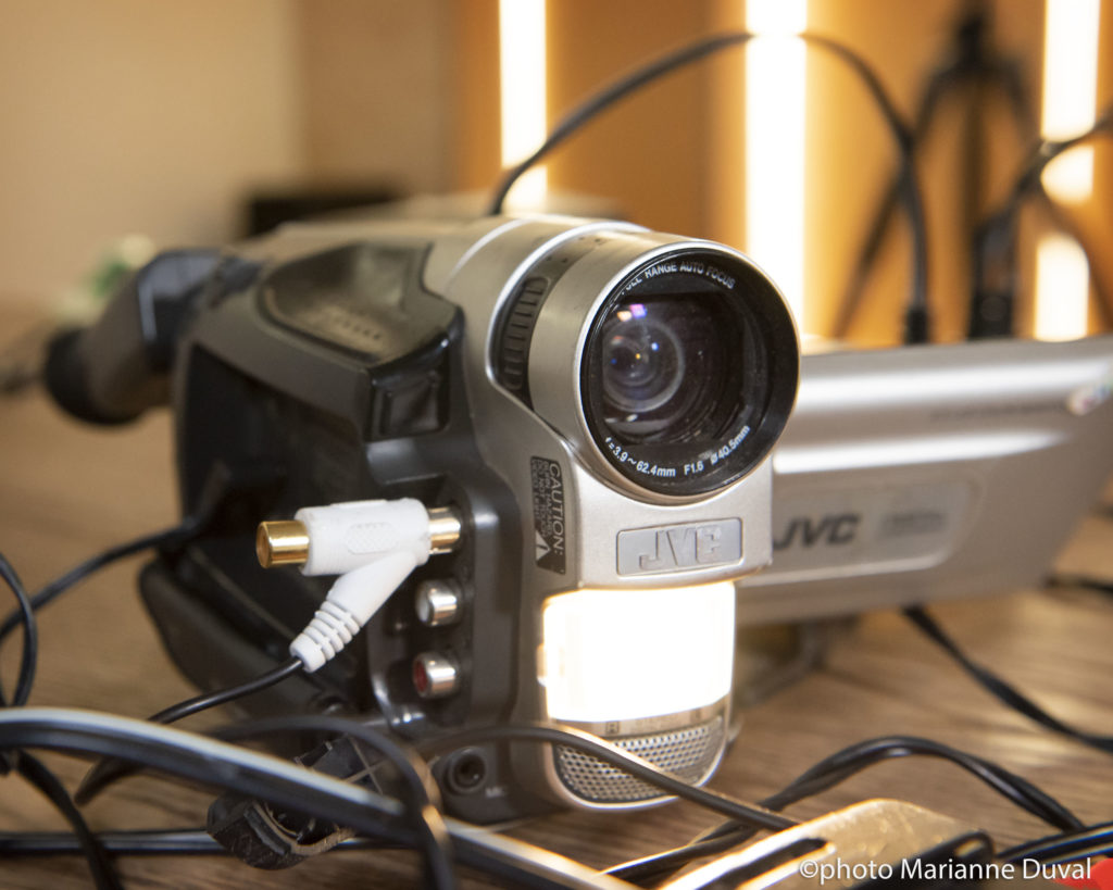 Close-up of a camcorder on a table with wires plugged into it and around it, and its display monitor open and tilted. / Gros plan d’un caméscope sur une table, le moniteur ouvert et incliné, entouré de fils don’t l’un y est branché.