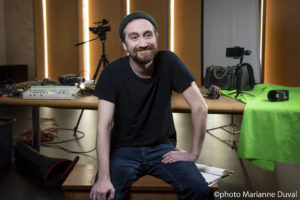 A smiling bearded White man sits, surrounded by camera equipment. / Un homme Blanc barbu souriant est assis, entouré d’équipement cinématographique.