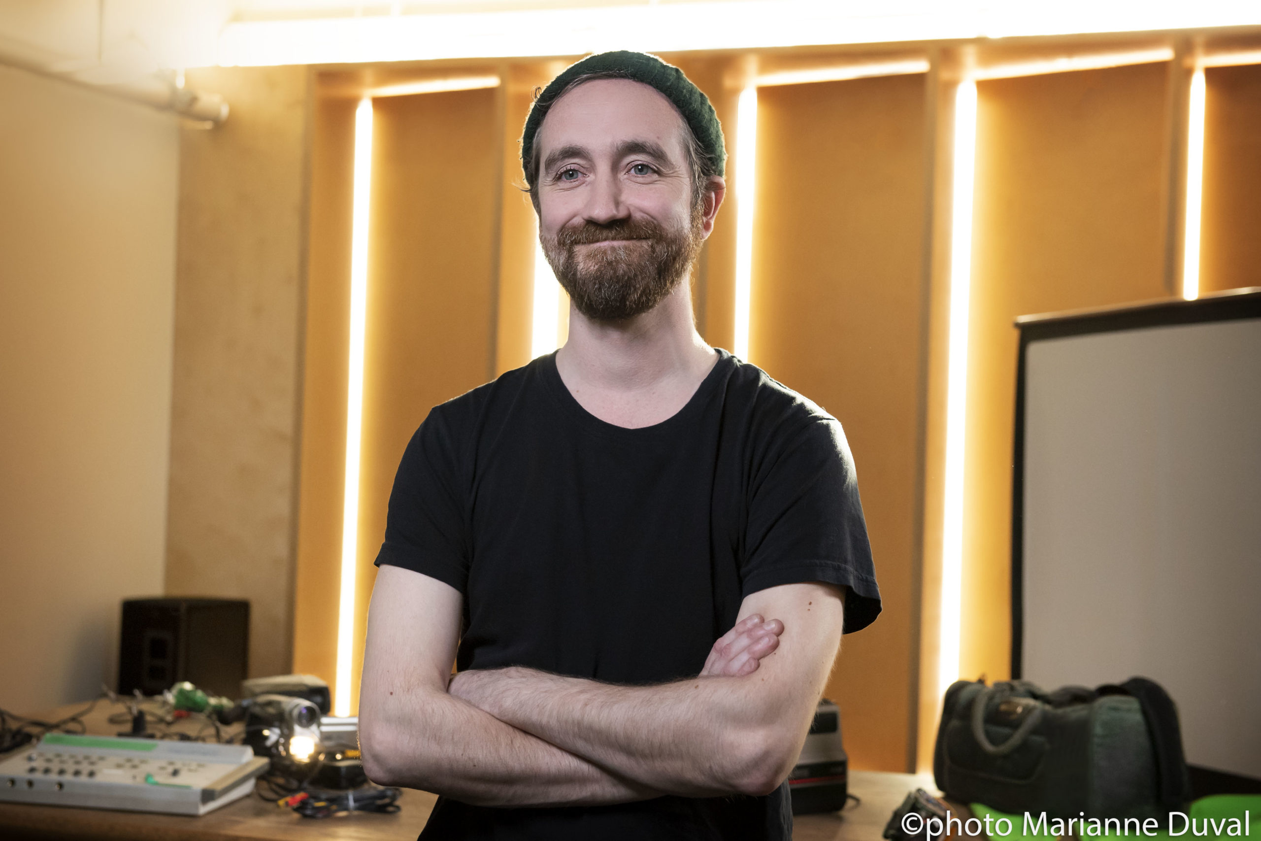 A smiling White bearded man with his arms crossed stands in a room scattered with camera equipment. / Un homme Blanc barbu souriant, les bras croisés, avec de l’équipement cinématographique éparpillé dans la salle.