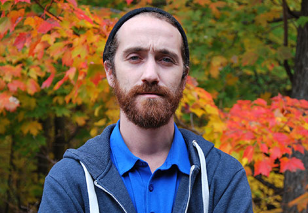 A White bearded man in front of autumn leaves looks at the camera. / Un homme Blanc barbu devant des feuilles d’automne regarde l’appareil photo.
