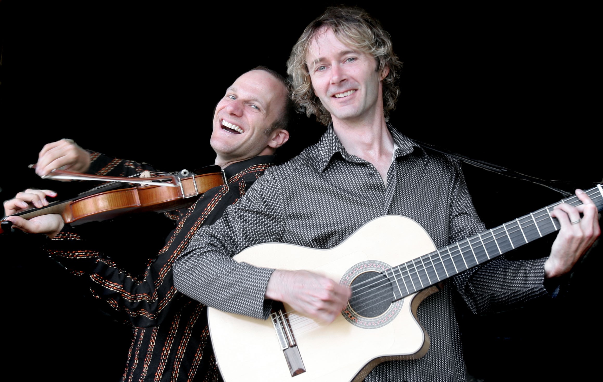 Two men lean their backs against one another, smiling as they play the violin and the flamenco guitar. / Deux hommes sourient et jouent le violon et la guitare flamenca dos à dos.
