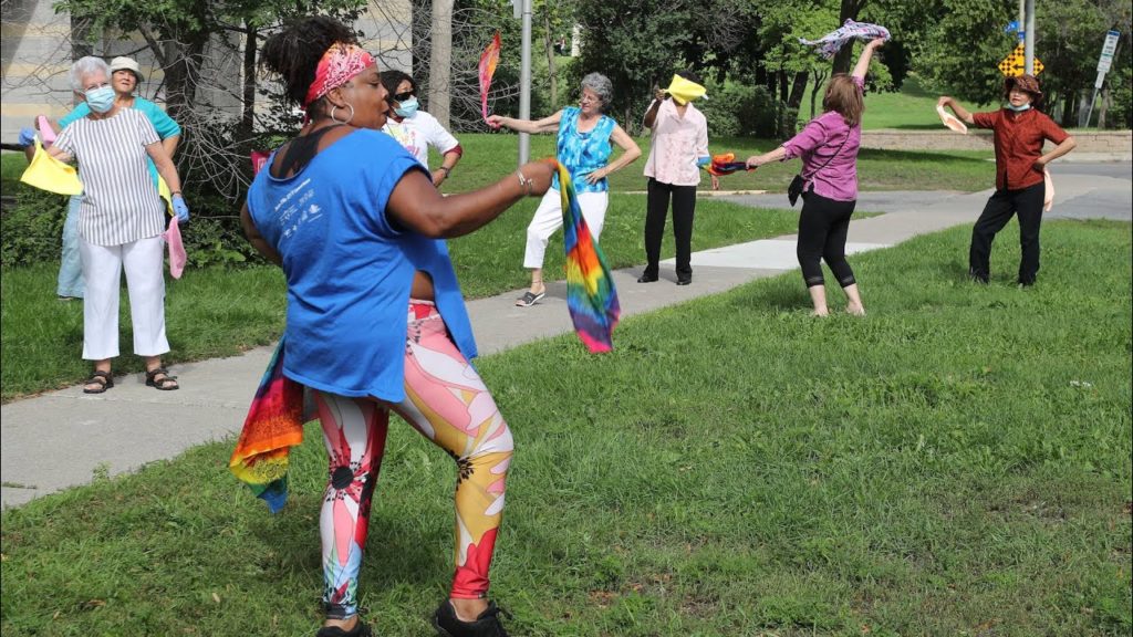 A Black woman demonstrates Afro Caribbean dance outside to seniors as they all wave colourful fabric around. / Une femme Noire fait une démonstration dehors de danse afro-antillaise et leur montre comment secouer leur tissus colorés pour des séniors.