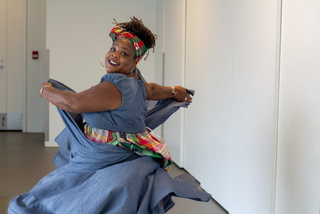 A smiling Black woman holds two ends of her denim-blue and twirls towards the camera. / Une femme Noire sourit et tient deux morceaux de sa robe bleu-jean et fait une pirouette vers la caméra.