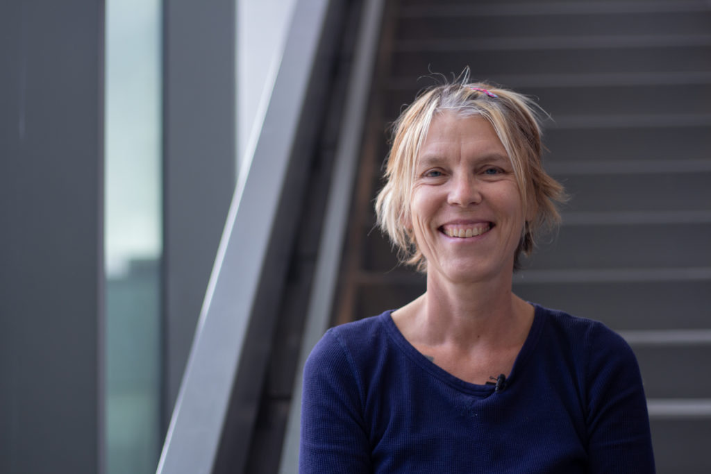 A smiling White woman with short blonde hair and blue eyes. / Une femme Blanche souriante aux cheveux blonds et courts et aux yeux bleus.
