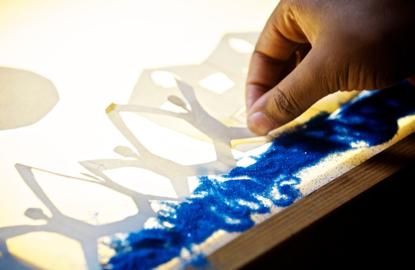 A hand places a white cutout chain of people next to a white cutout of a house and above a thick horizontal row of dark blue glitter on a lit up pane of glass. / Une main place une chaîne blanche découpée de personnes à côté d’une maison blanche découpée au dessus d’une rangée épaisse horizontale de paillettes bleues foncées sur une vitre illuminée.