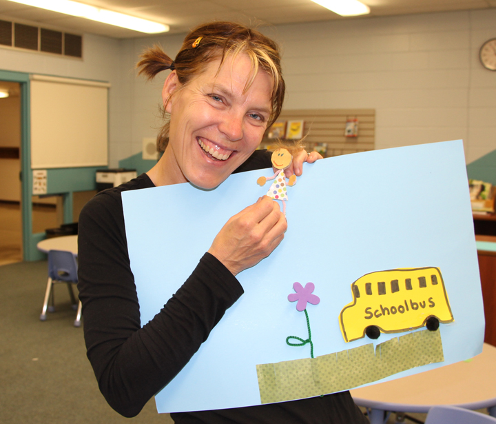 A smiling White woman in a classroom holds up a pale blue poster board with a flower, school bus and little girl glued on. / Une femme Blanche souriante dans une classe tient un poster bleu pâle avec collé dessus un autobus scolaire, une fleur et une petite fille.
