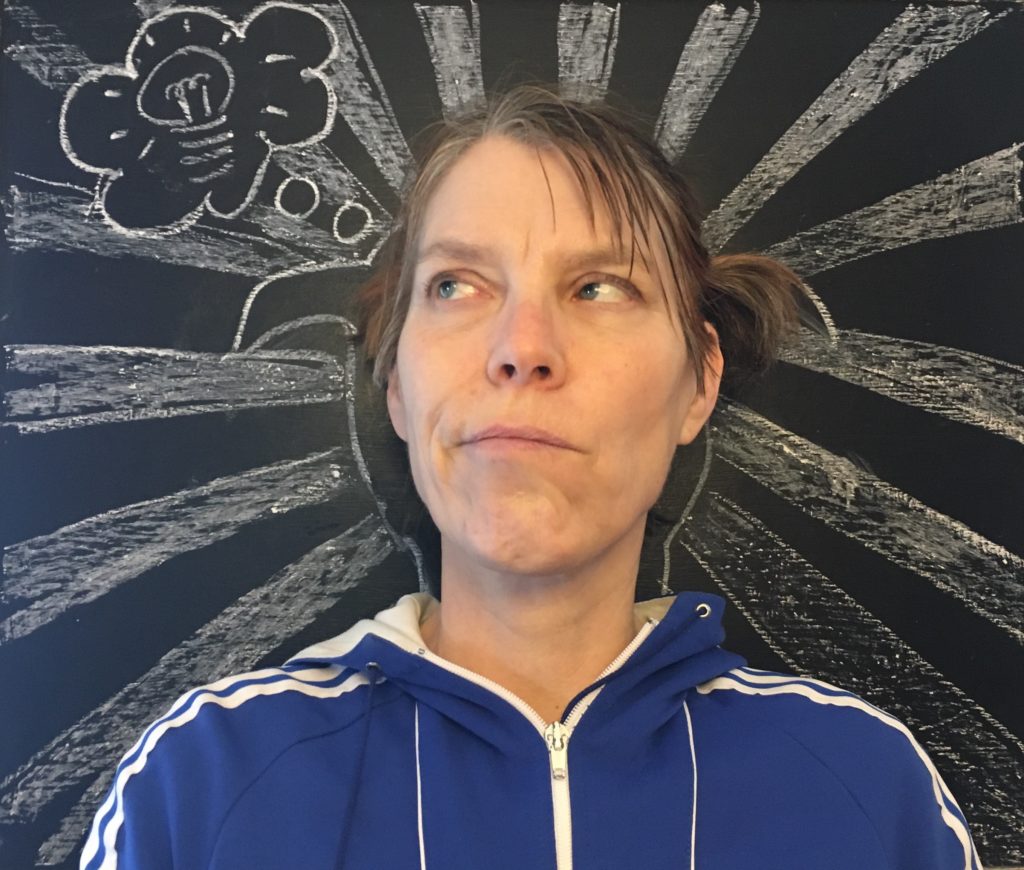 A White woman looks up pensively in front of a chalkboard with an outline of her with thick rays shooting out and a thought bubble populated by a lit lightbulb drawn in white chalk. / Une femme Blanche regarde vers le ciel pensivement devant un tableau noir avec des rayons épais qui proviennent d’un tracé de sa tête et une bulle de pensée avec une ampoule illuminée à l’intérieur dessiné avec de la craie blanche.
