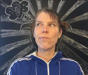 A White woman looks up pensively in front of a chalkboard with an outline of her with thick rays shooting out and a thought bubble populated by a lit lightbulb drawn in white chalk. / Une femme Blanche regarde vers le ciel pensivement devant un tableau noir avec des rayons épais qui proviennent d’un tracé de sa tête et une bulle de pensée avec une ampoule illuminée à l’intérieur dessiné avec de la craie blanche.