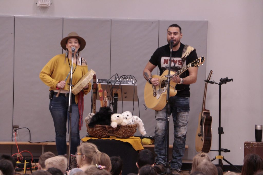 An Indigenous duo performs music for an audience of children seated in a gymnasium; both sing into microphones, the woman also plays the traditional hand drum and the man plays guitar. / Un duo Autochtone joue de la musique dans un gymnase pour des enfants assis parterre; la femme chante et joue du tambour à main traditionnel, l’homme chante et joue de la guitare.