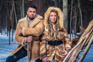 An Indigenous man and woman pose in a forest in the winter. / Un homme et femme autochtones posent dans une forêt en hiver.