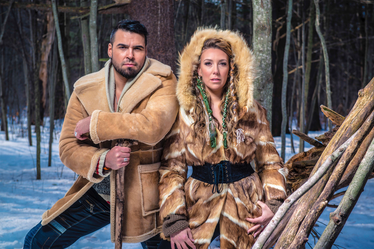 An Indigenous man and woman pose in a forest in the winter. / Un homme et femme autochtones posent dans une forêt en hiver.