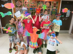 A group of children wear masks they have made and hold up their illustrations on sticks. / Un groupe d’enfants porte des masques qu’ils ont créés et montrent leurs illustrations sur des bâtons.