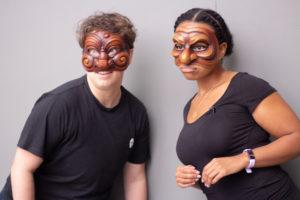 A White non-binary person and Black woman wear wooden Commedia Dell’Arte masks. / Une personne Blanche non-binaire et une femme Noire portent des masques en bois de la Commedia Dell’Arte.