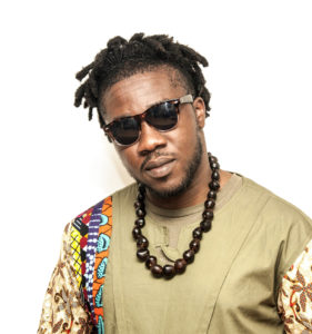 A Black man wears sunglasses and a large brown beaded necklace over a green shirt with multicoloured patterned sleeves. / Un homme Noir porte des lunettes de soleil et un grand collier marron perlé par dessus un chandail vert aux manches multicolores et à motifs.