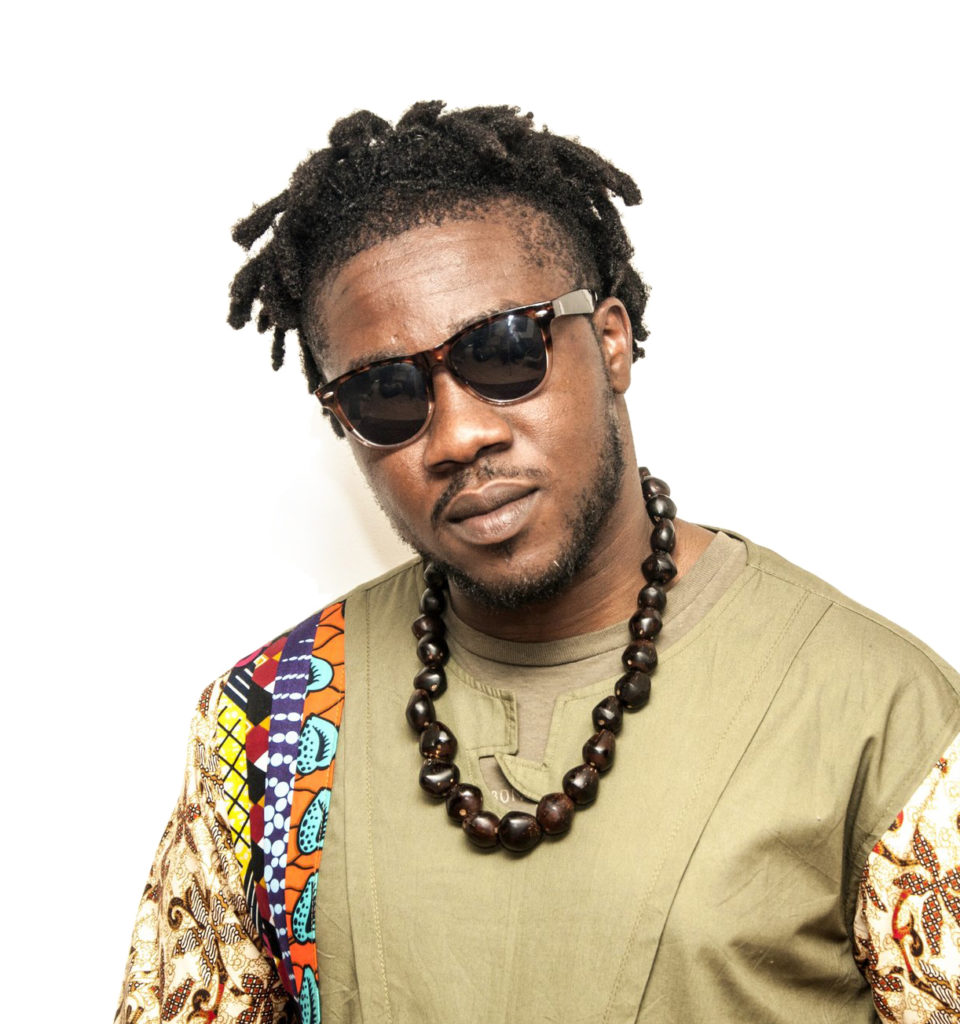 A Black man wears sunglasses and a large brown beaded necklace over a green shirt with multicoloured patterned sleeves. / Un homme Noir porte des lunettes de soleil et un grand collier marron perlé par dessus un chandail vert aux manches multicolores et à motifs.