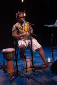 A Black man’s legs on a stage next to a hand drum. / Les jambes d’un homme Noir à côté d’un tambour à main sur une scène.
