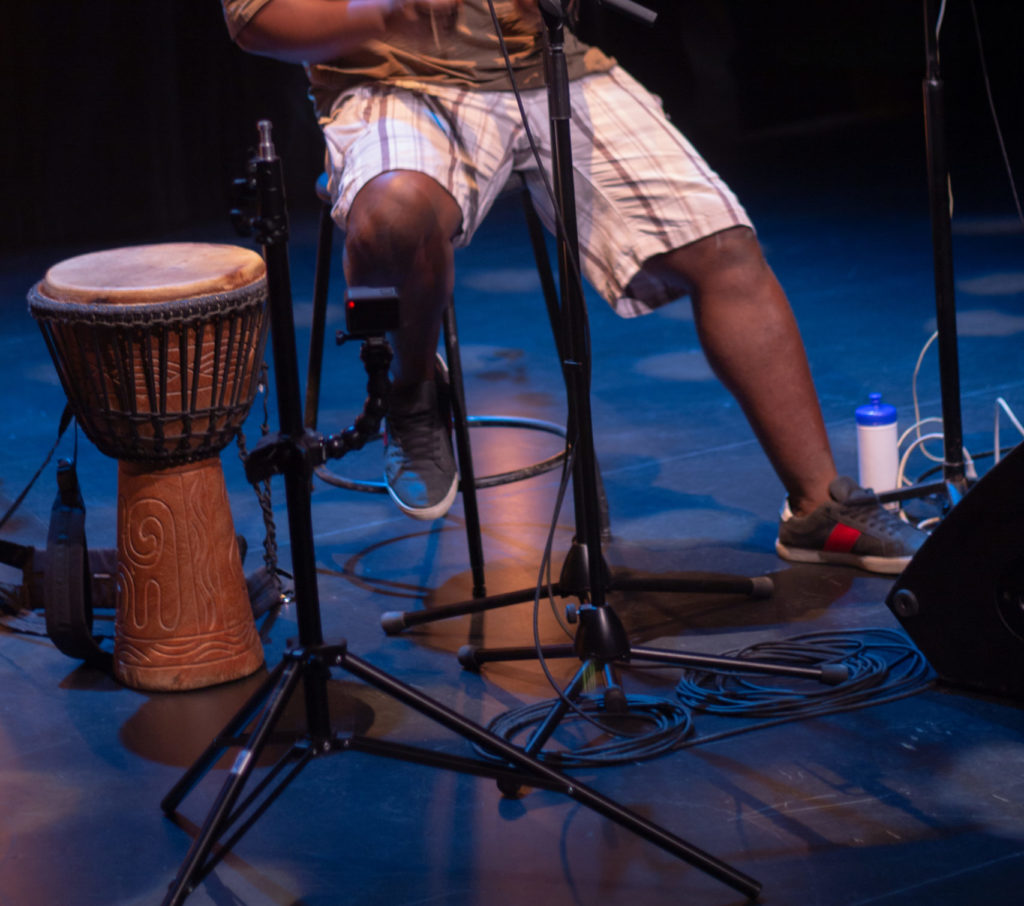 A Black man’s legs on a stage next to a hand drum. / Les jambes d’un homme Noir à côté d’un tambour à main sur une scène.