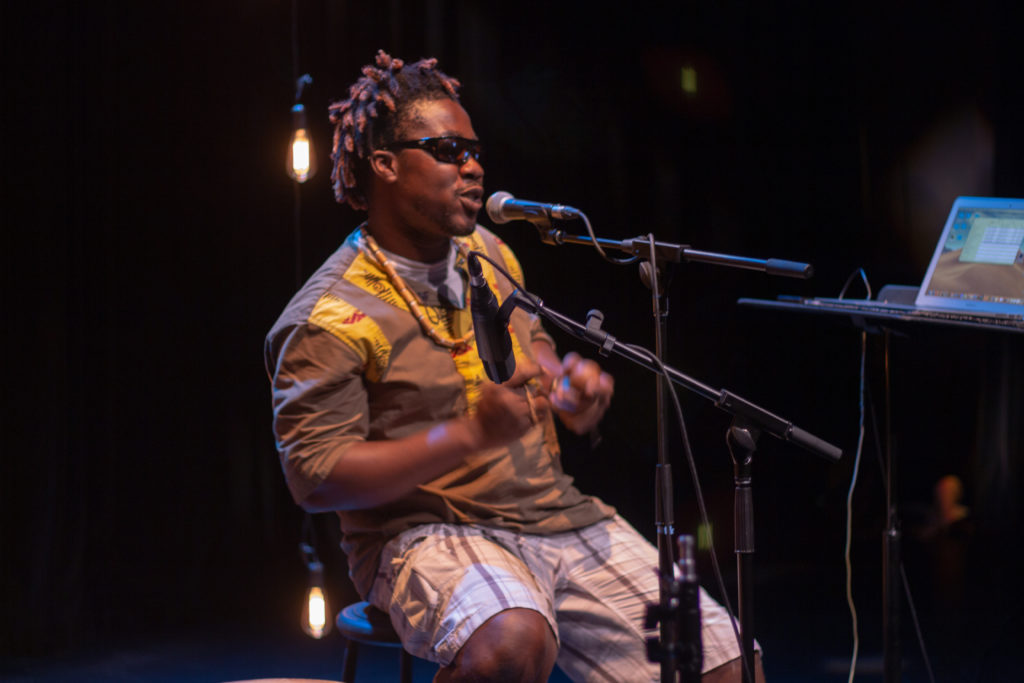 A seated Black man with sunglasses on sings into a microphone. / Un homme Noir assis portant des lunettes de soleil chante dans un microphone.